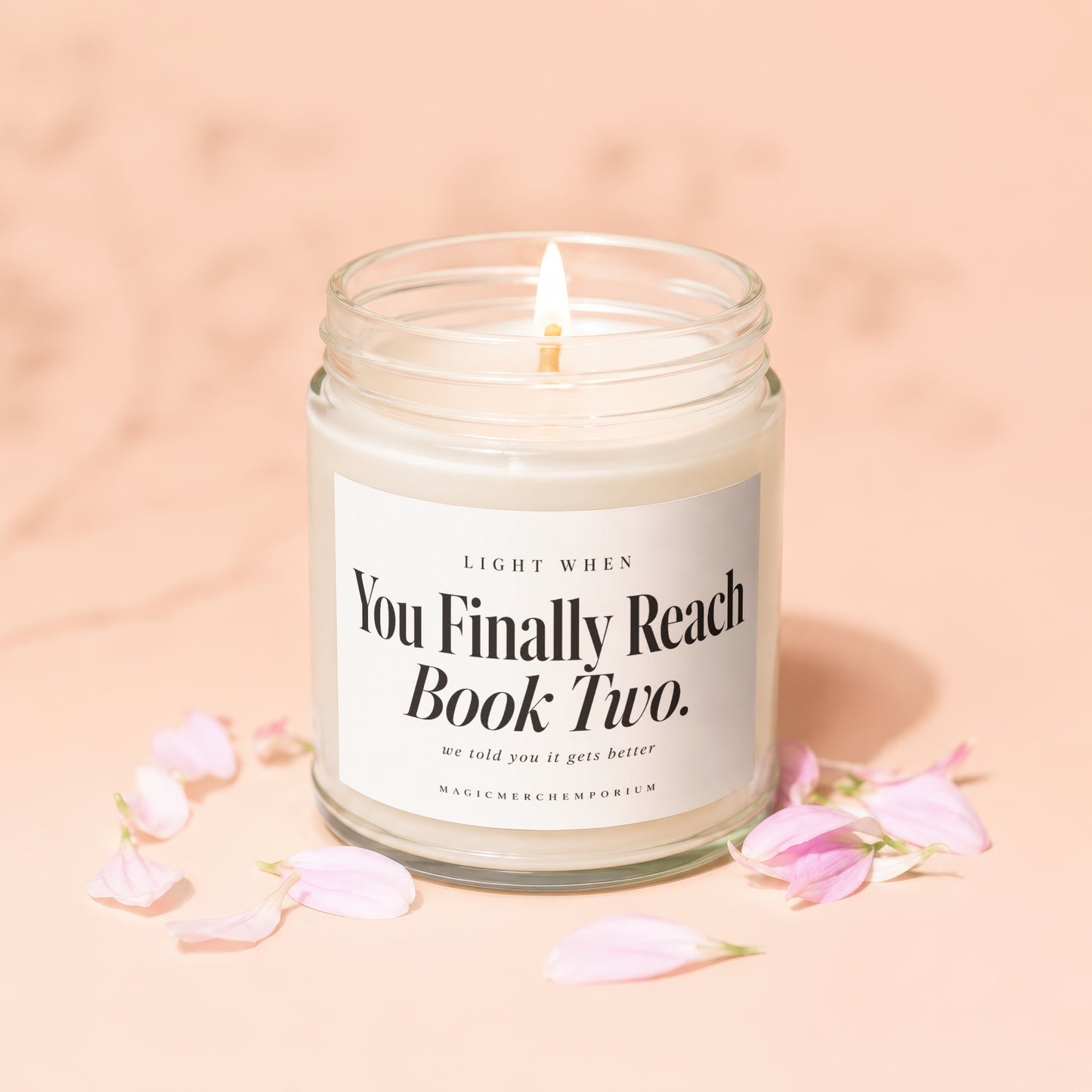 A lit candle in a glass jar with a white label reading You Finally Reach Book Two against a peach background scattered with pink petals.