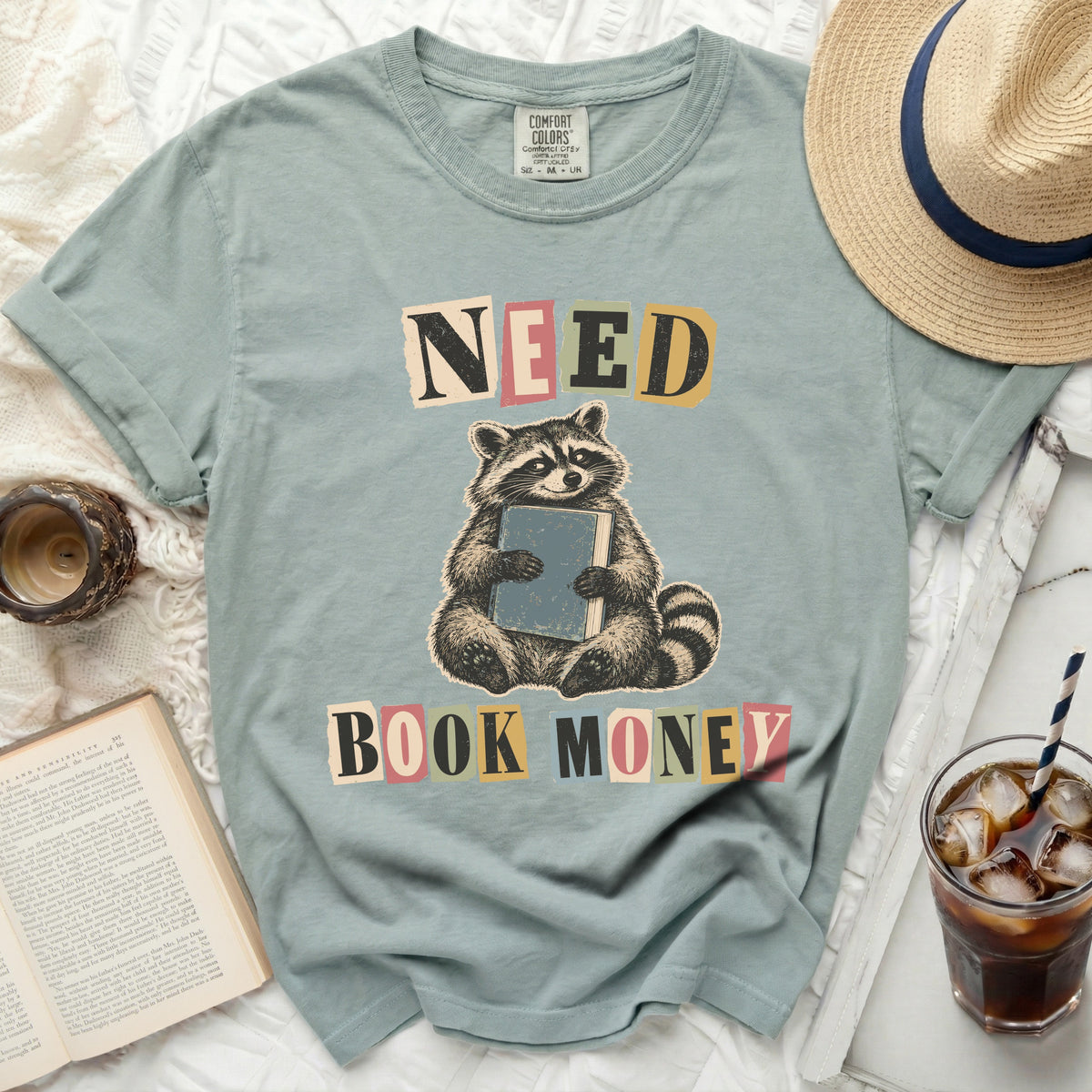 Light blue t-shirt with a graphic of a raccoon holding a book and the words "NEED BOOK MONEY".