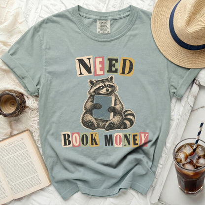 Light blue t-shirt with a graphic of a raccoon holding a book and the words "NEED BOOK MONEY".