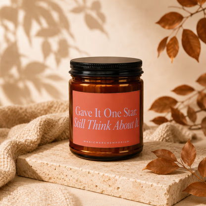 Amber candle with black lid and coral label reading "Gave It One Star. Still Think About It."