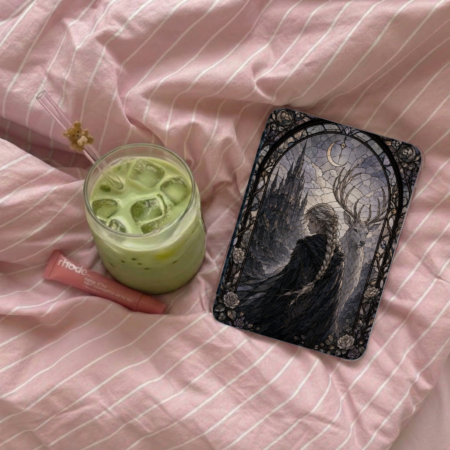 Glass of matcha with ice, a lip balm, and a phone case on pink striped sheets.