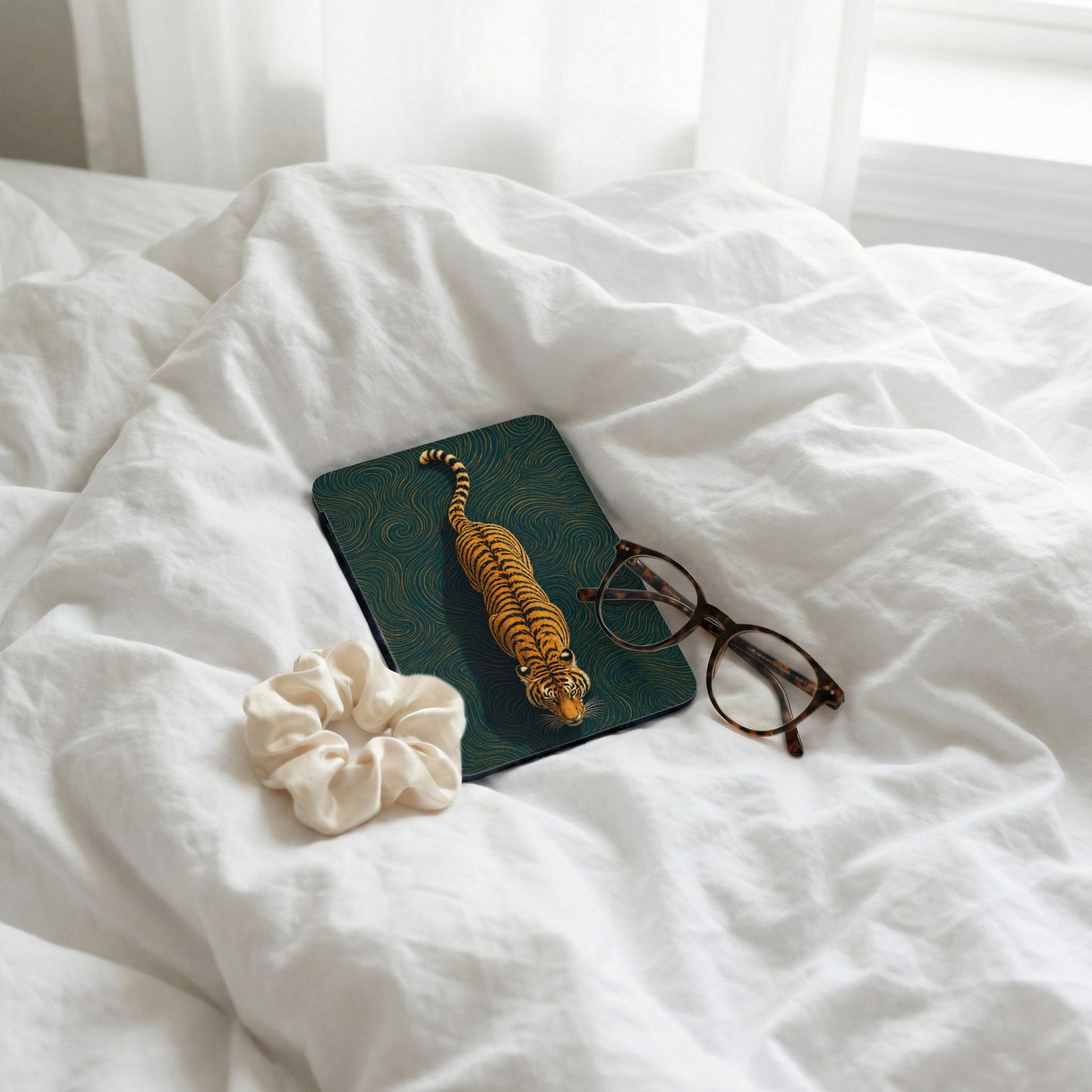 Mousepad with tiger design next to glasses and scrunchie on wrinkled white sheets.