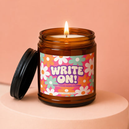 Amber candle with lit wick and floral label saying "WRITE ON! KEEP THOSE PAGES COMING".