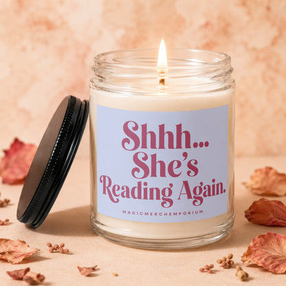 Glass candle with a lit wick and black lid, reading "Shhh... She's Reading Again."