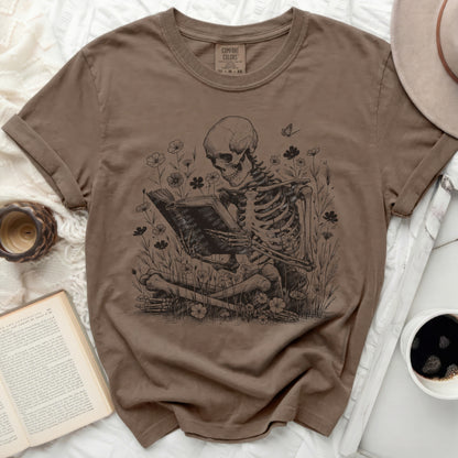 Brown t-shirt with skeleton reading a book amidst flowers and butterflies.
