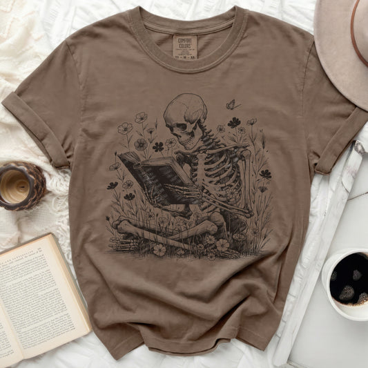 Brown t-shirt with skeleton reading a book amidst flowers and butterflies.