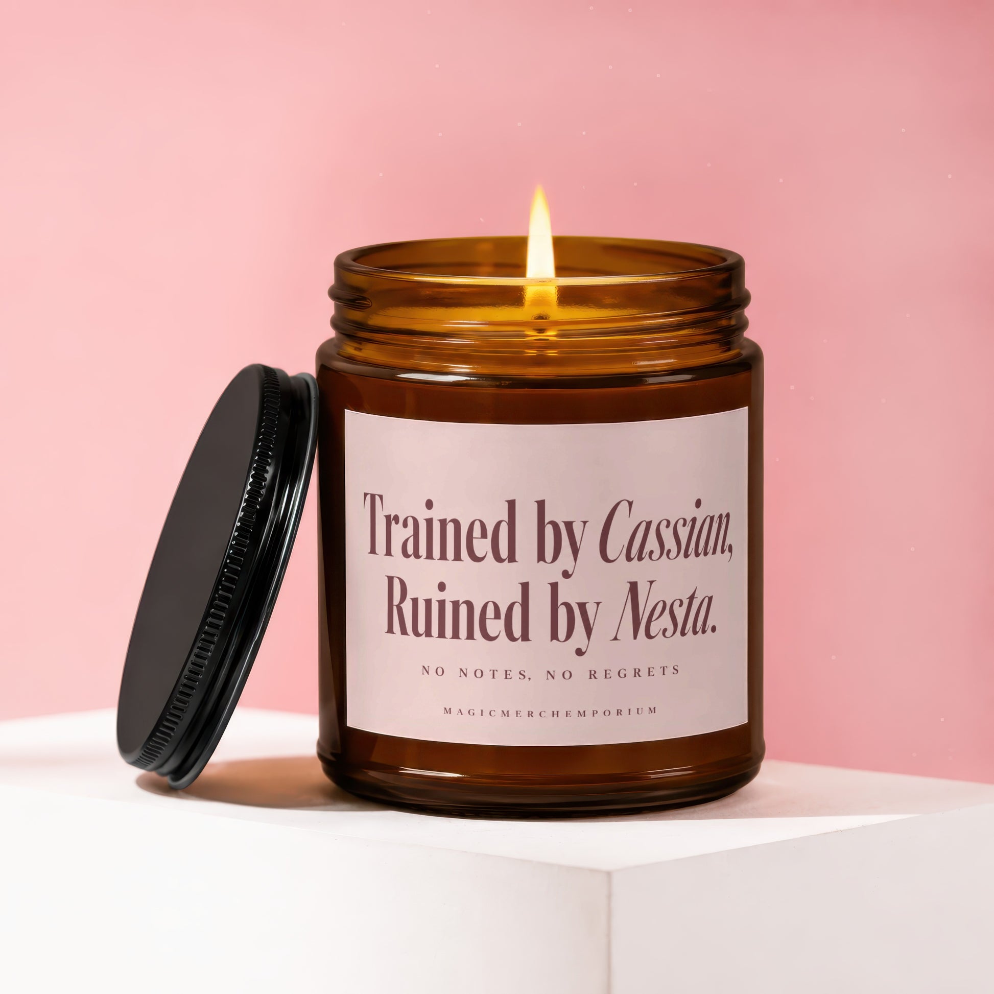Amber glass candle with lit wick and black lid. Label reads: "Trained by Cassian, Ruined by Nesta."