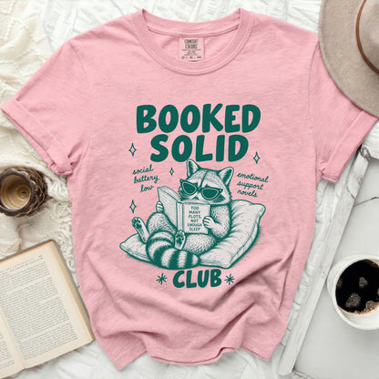 Pink t-shirt with raccoon reading "Too Many Plots, Not Enough Sleep" and "Booked Solid Club" text.