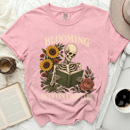 Pink t-shirt with skeleton reading a book surrounded by sunflowers and flowers, text reads "Blooming Between Chapters".