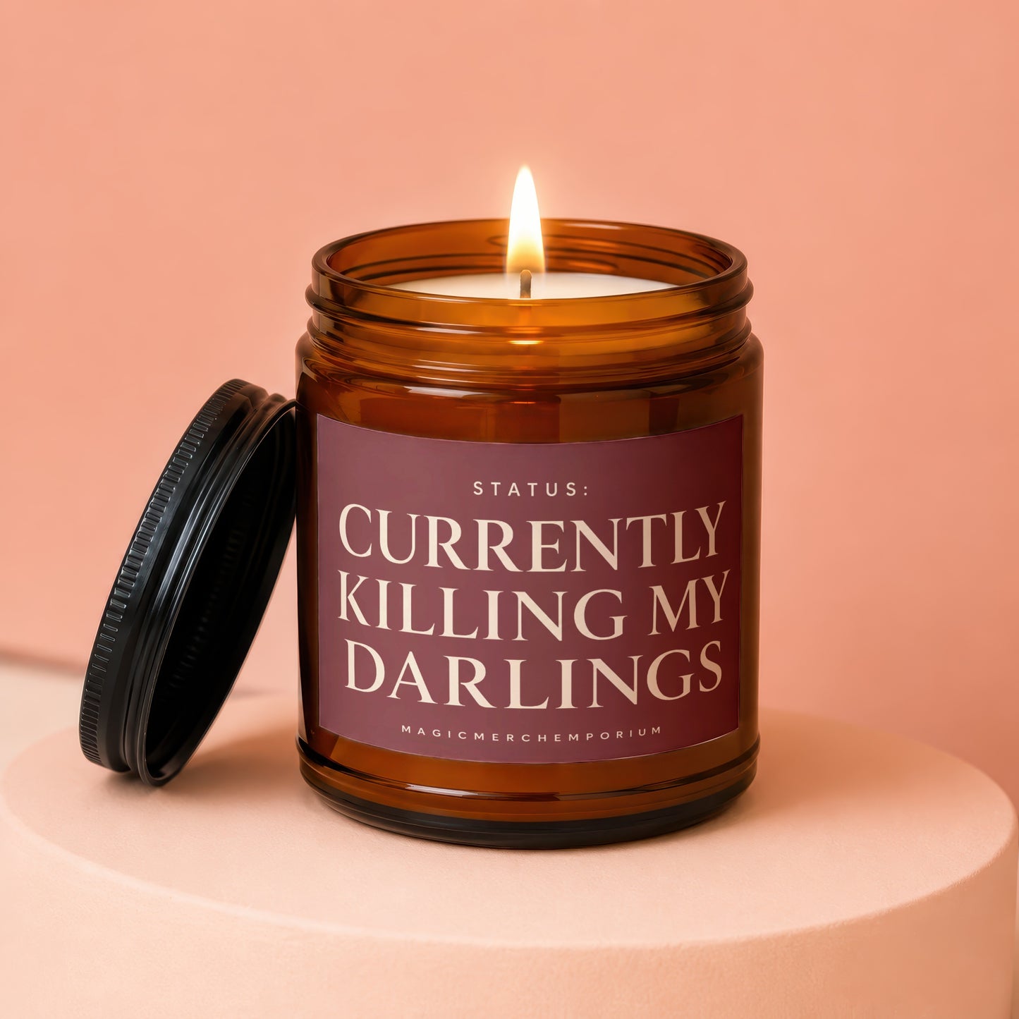 Amber glass candle with burning wick and label reading "STATUS: CURRENTLY KILLING MY DARLINGS."