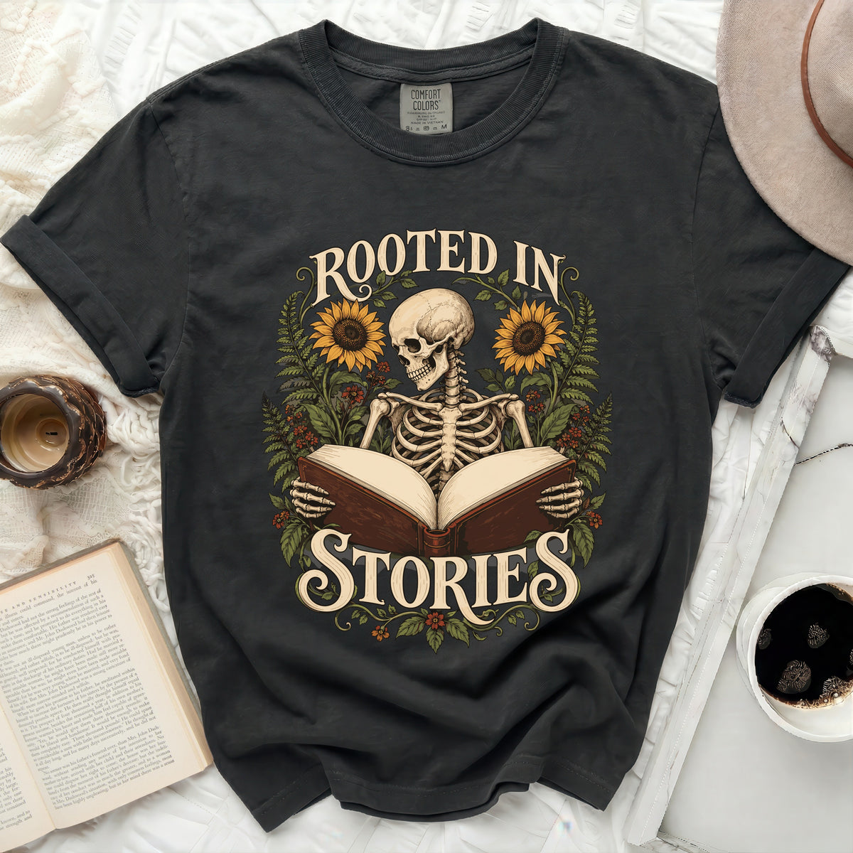 Skeleton reading an open book with sunflowers and foliage, text "ROOTED IN STORIES".