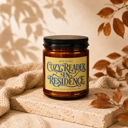 Amber candle with black lid, "Cozy Reader" label, near knitted fabric and fall leaves.