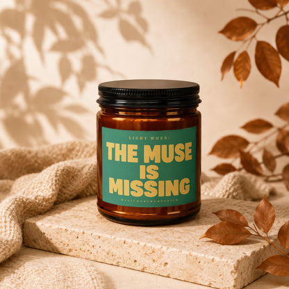 Amber candle with black lid, green label reads "The Muse is Missing".