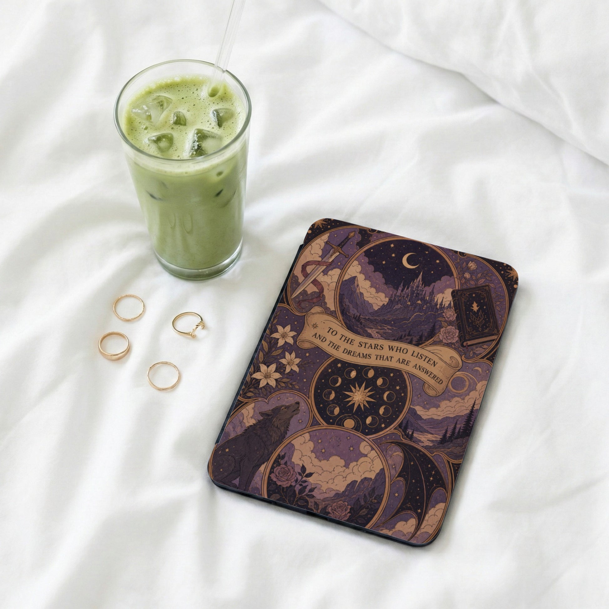 Book with enchanting mystical art, featuring a wolf, castle, and text, next to iced matcha and rings.