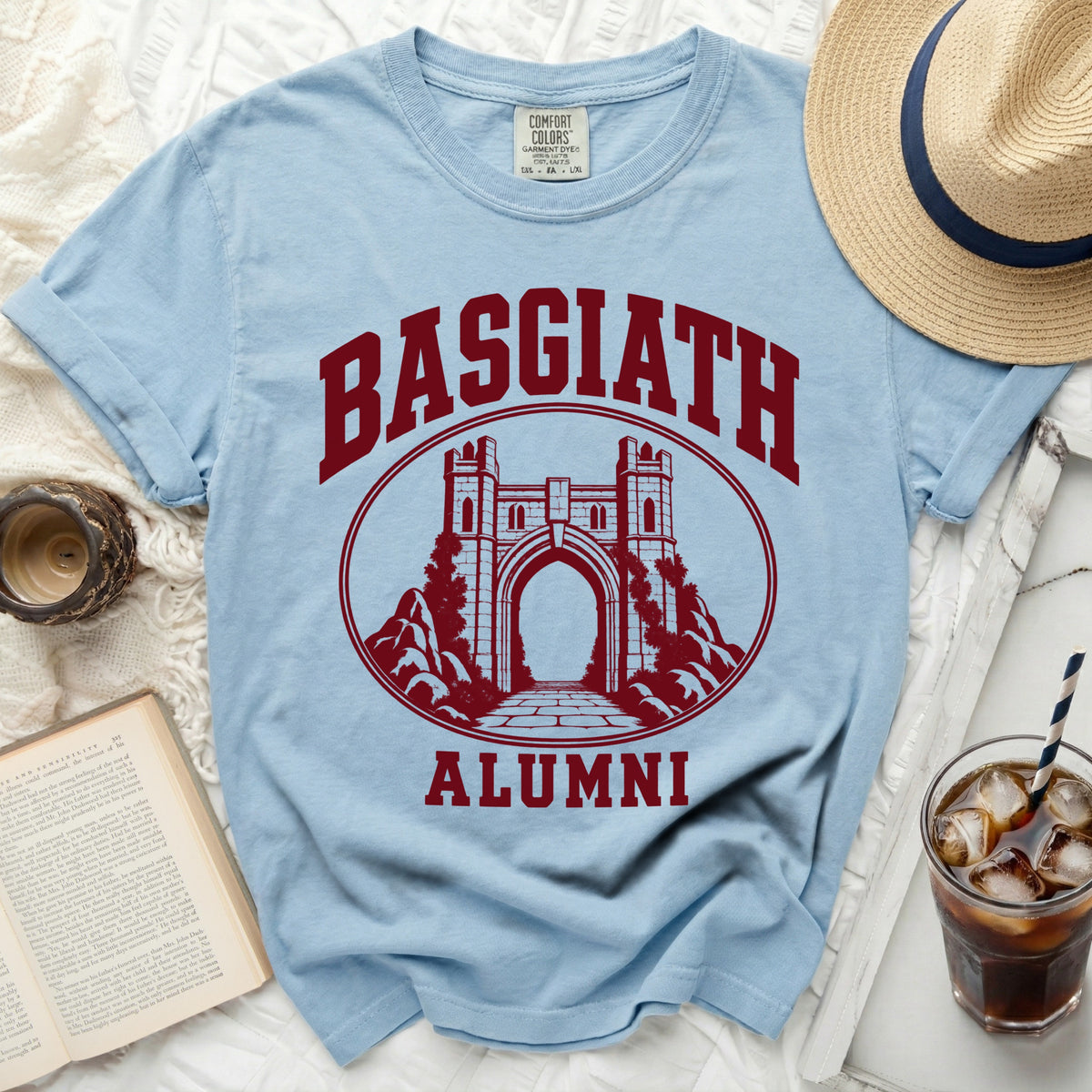 Light blue crewneck t-shirt featuring a maroon Basgiath Alumni castle graphic, surrounded by a straw hat, iced drink, and book.