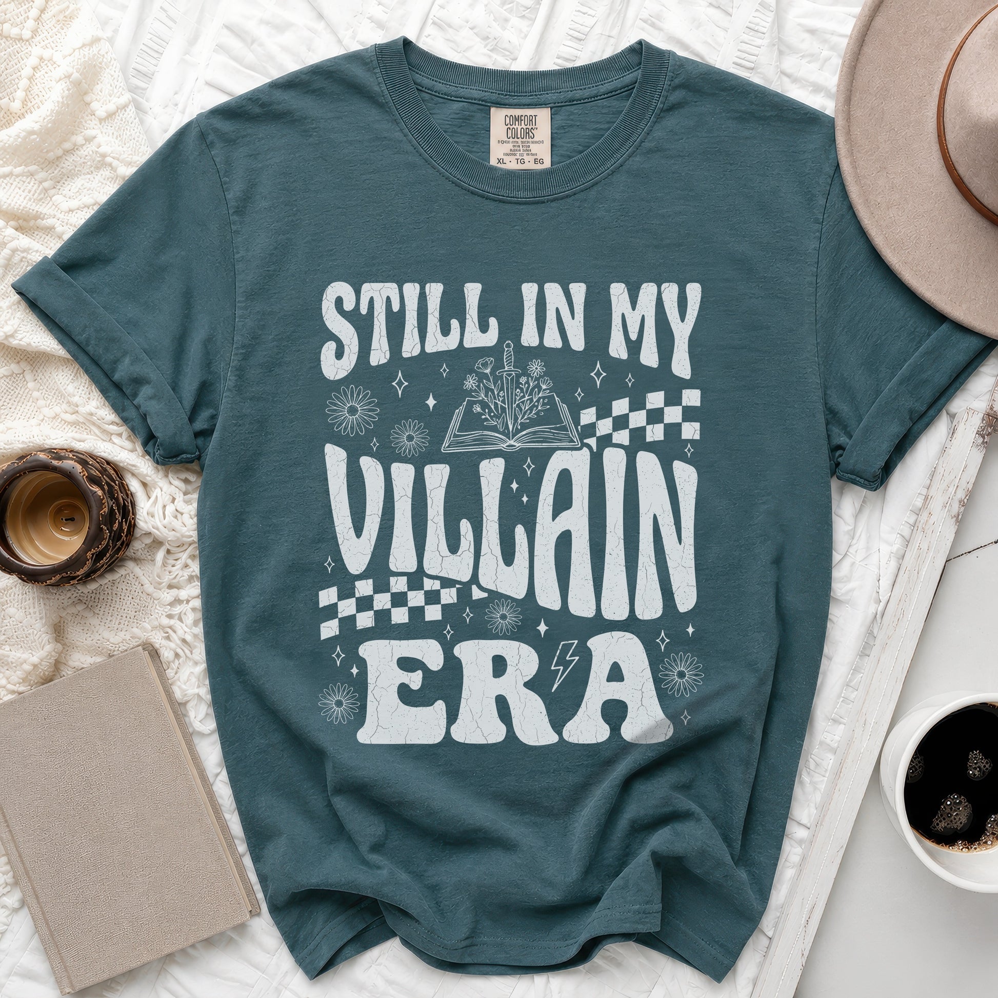 Teal t-shirt with white "Still in My Villain Era" text, book, sword, and flowers.