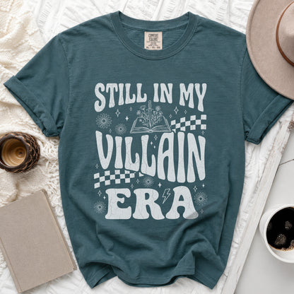 Teal t-shirt with white "Still in My Villain Era" text, book, sword, and flowers.