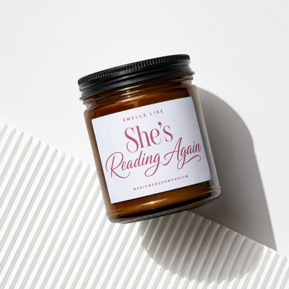 Brown glass candle with white label reading "She's Reading Again".