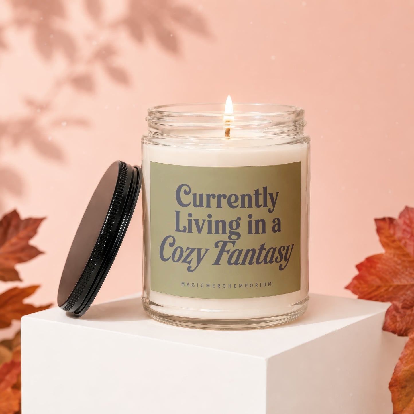 Glass candle with lit wick, cap removed, reading "Currently Living in a Cozy Fantasy".