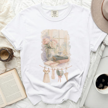 White t-shirt with watercolor print of clothes on a line, books, and flowers.