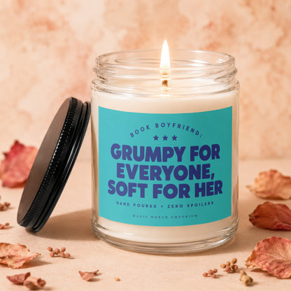 Candle with label "Grumpy for everyone, soft for her" on a textured surface with dried leaves.