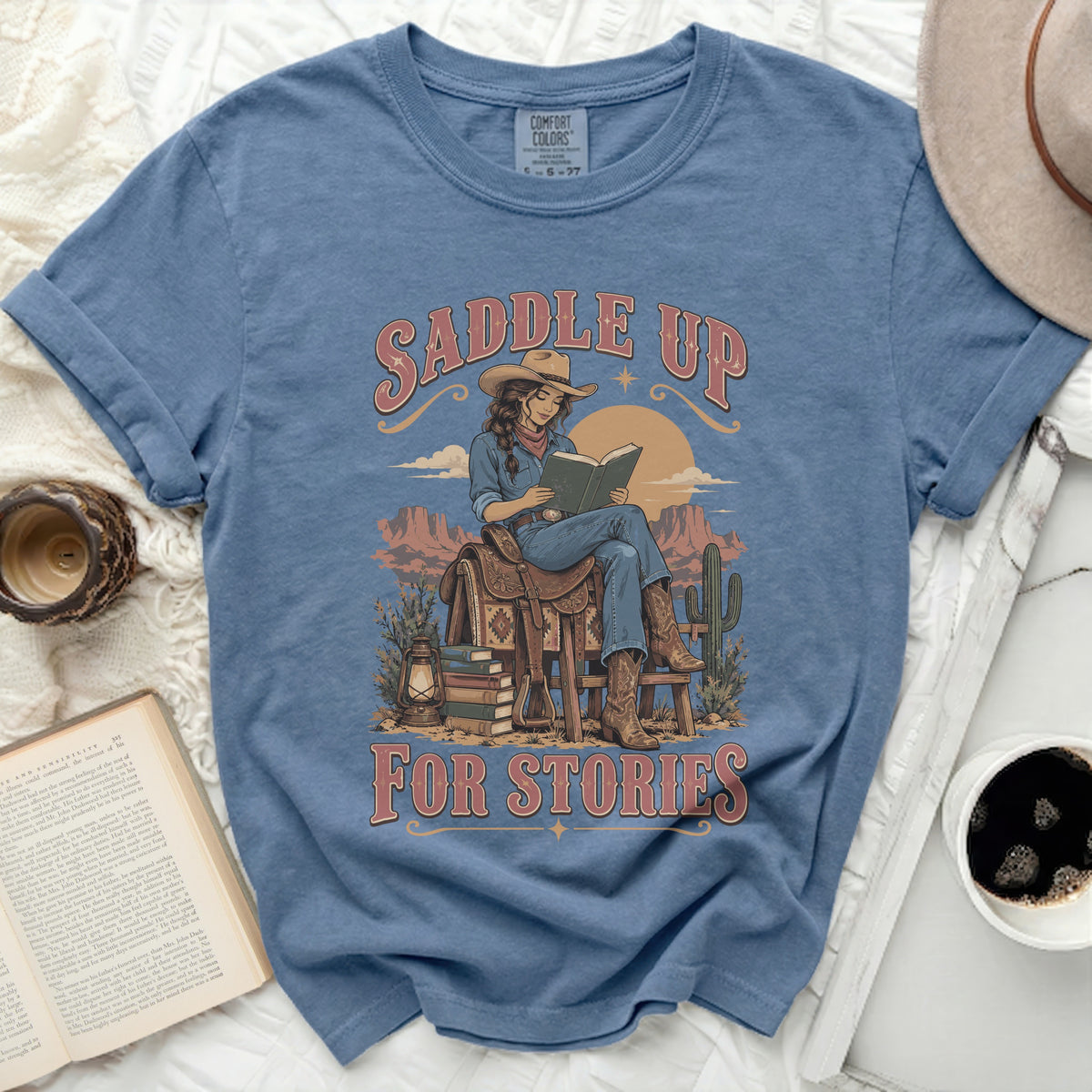 Blue t-shirt with "Saddle Up For Stories" text and cowgirl reading on a saddle.