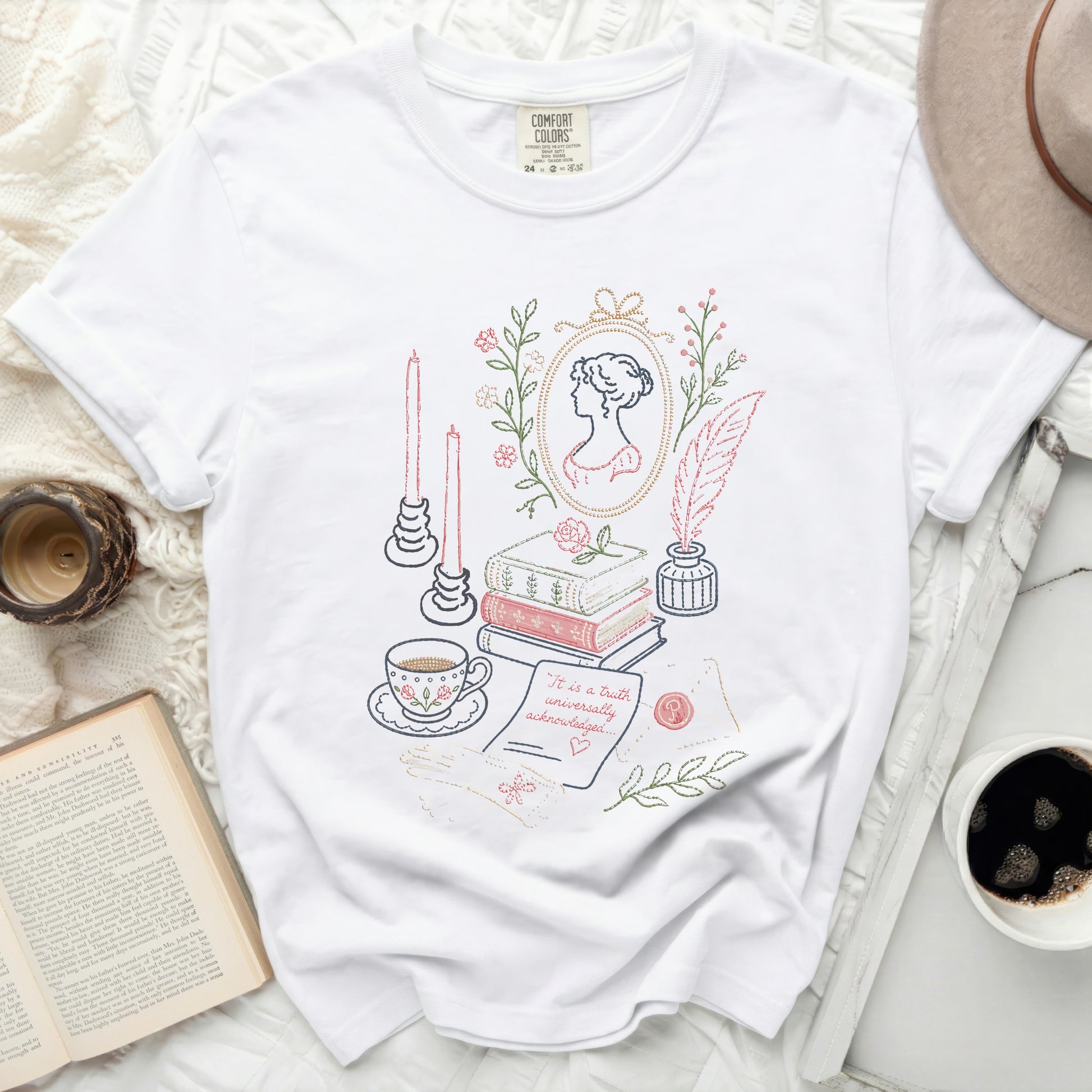 White t-shirt with embroidered scene: cameo, books, teacup, quill, and candles.