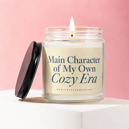 Glass candle with a lit wick and a black lid, featuring text "Main Character of My Own Cozy Era."