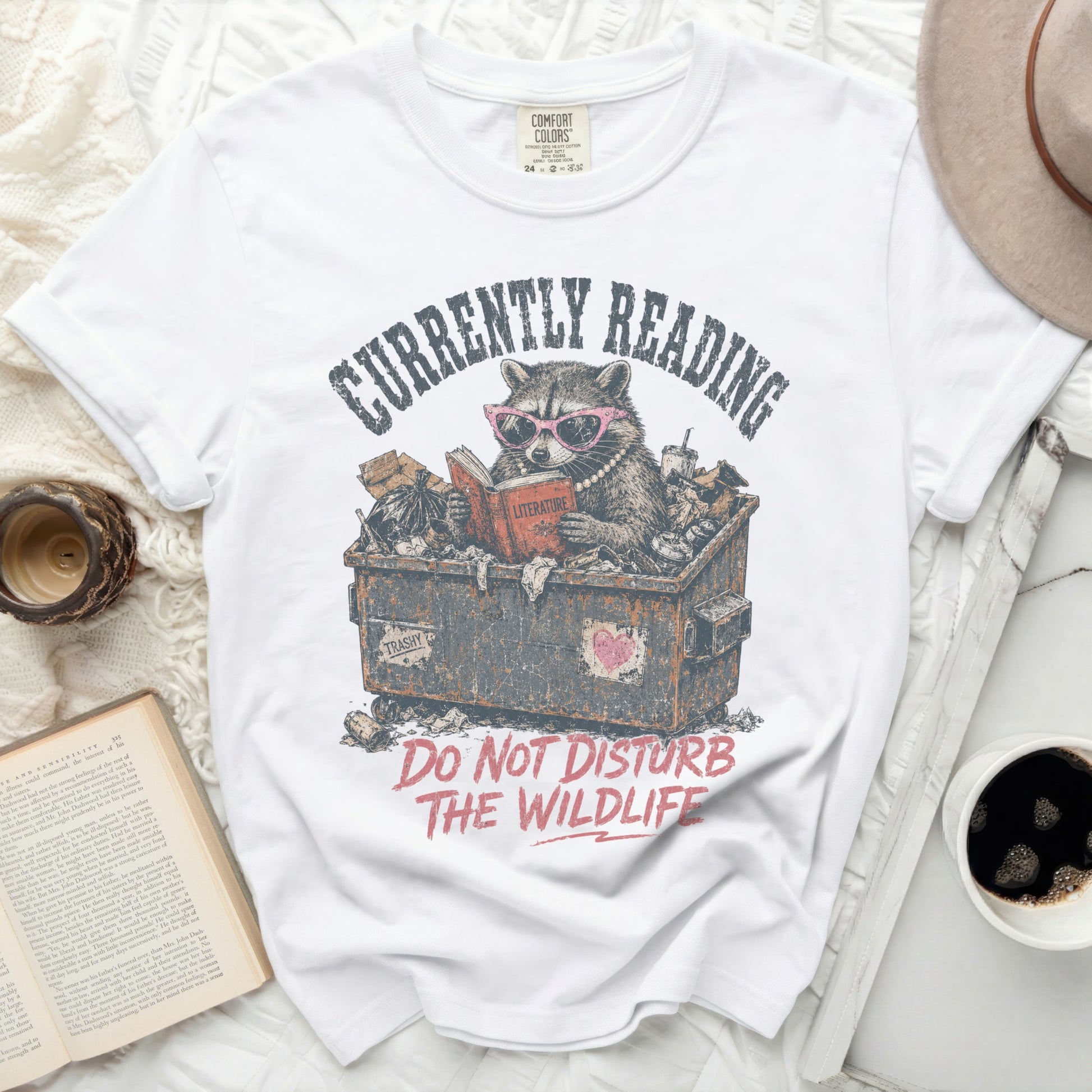 White t-shirt with graphic of raccoon reading in trash bin: "Currently Reading Literature".