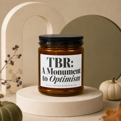 Amber jar candle with black lid reading "TBR: A Monument to Optimism"