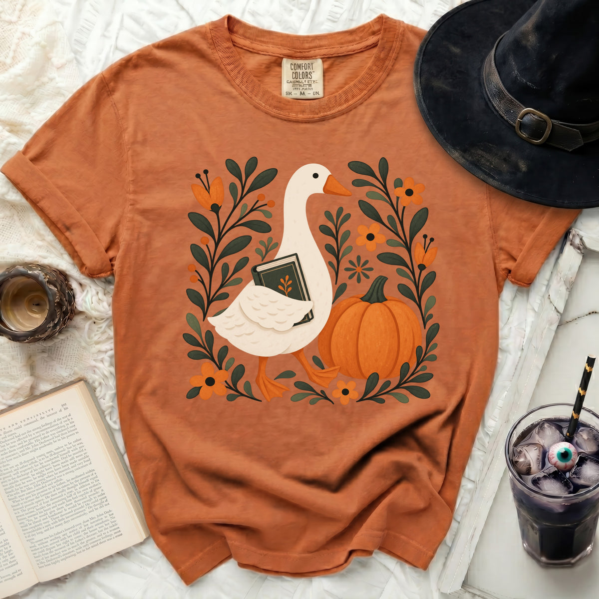 Orange t-shirt with a goose holding a book, surrounded by flowers and a pumpkin.