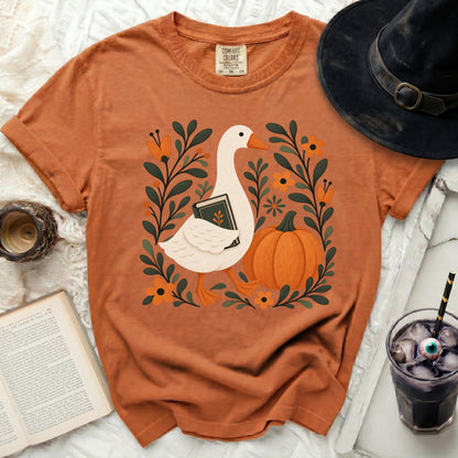 Orange t-shirt with a goose holding a book, surrounded by flowers and a pumpkin.