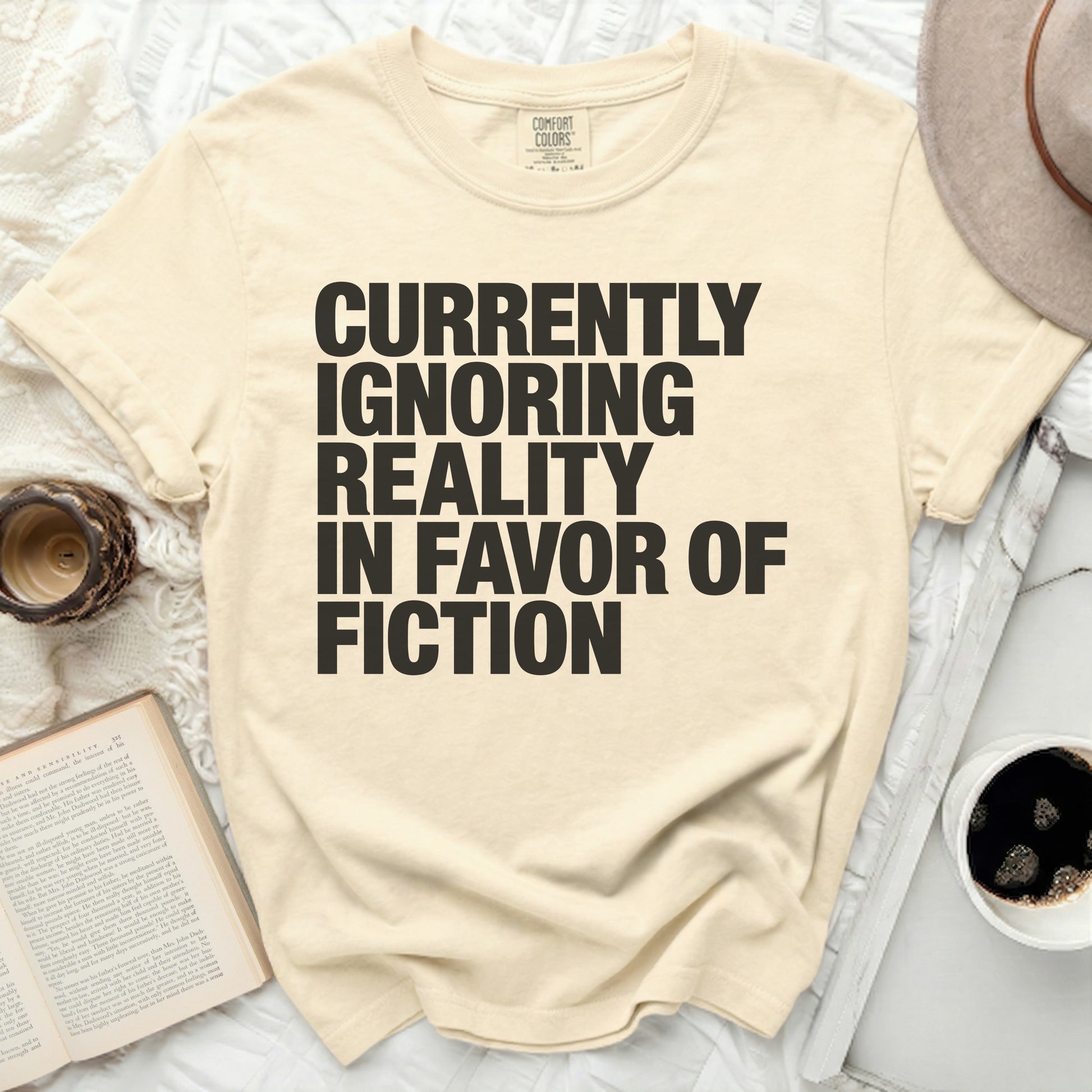 Cream t-shirt with black text reading "Currently Ignoring Reality In Favor Of Fiction".