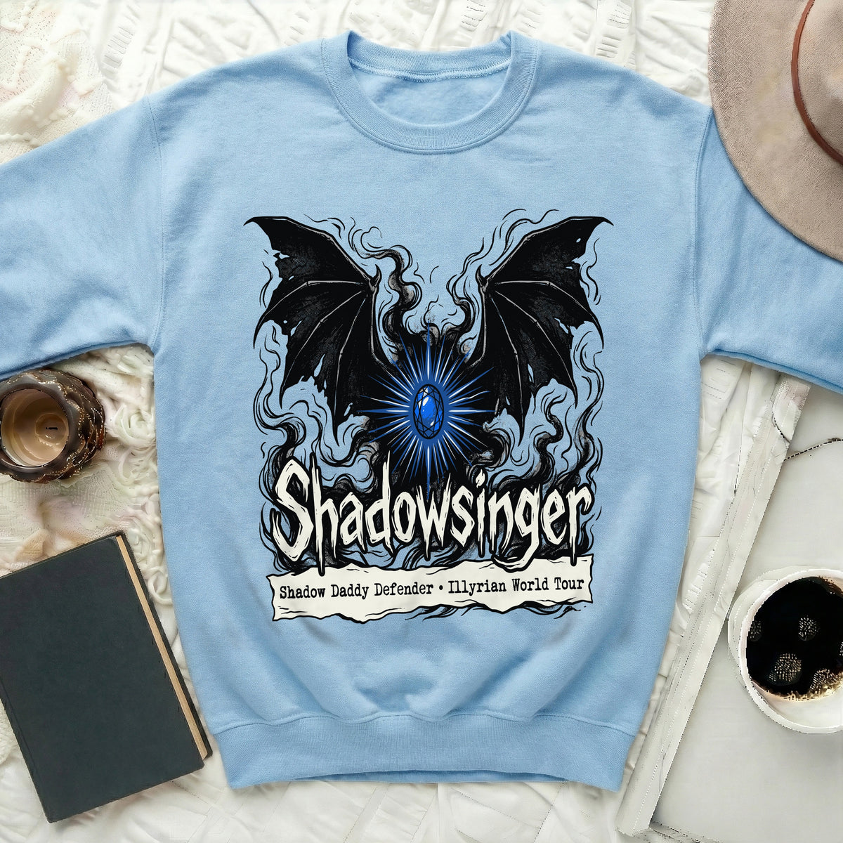 Light blue crewneck sweatshirt featuring a graphic with bat wings, a glowing blue jewel, and Shadowsinger text.