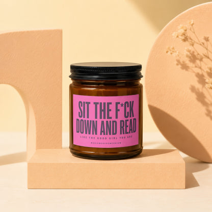 Candle with pink label reading "SIT THE F*CK DOWN AND READ LIKE THE GOOD GIRL YOU ARE".