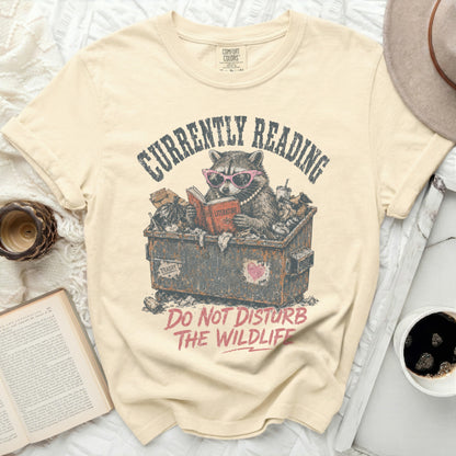 Cream t-shirt with raccoon reading in trash bin graphic and text.