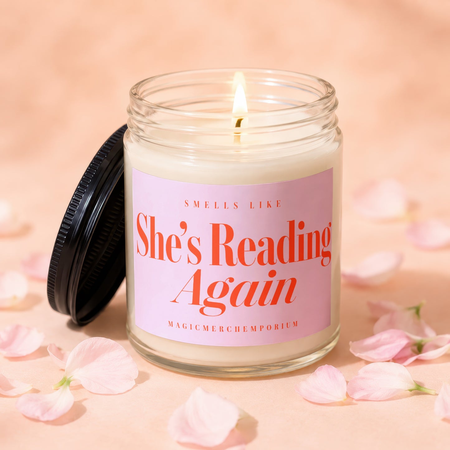 Candle with lit wick and pink label that says "She's Reading Again".