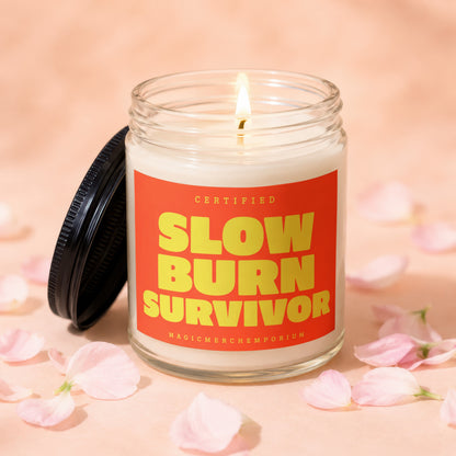 Glass candle with burning wick and bright orange label reading "Certified Slow Burn Survivor".