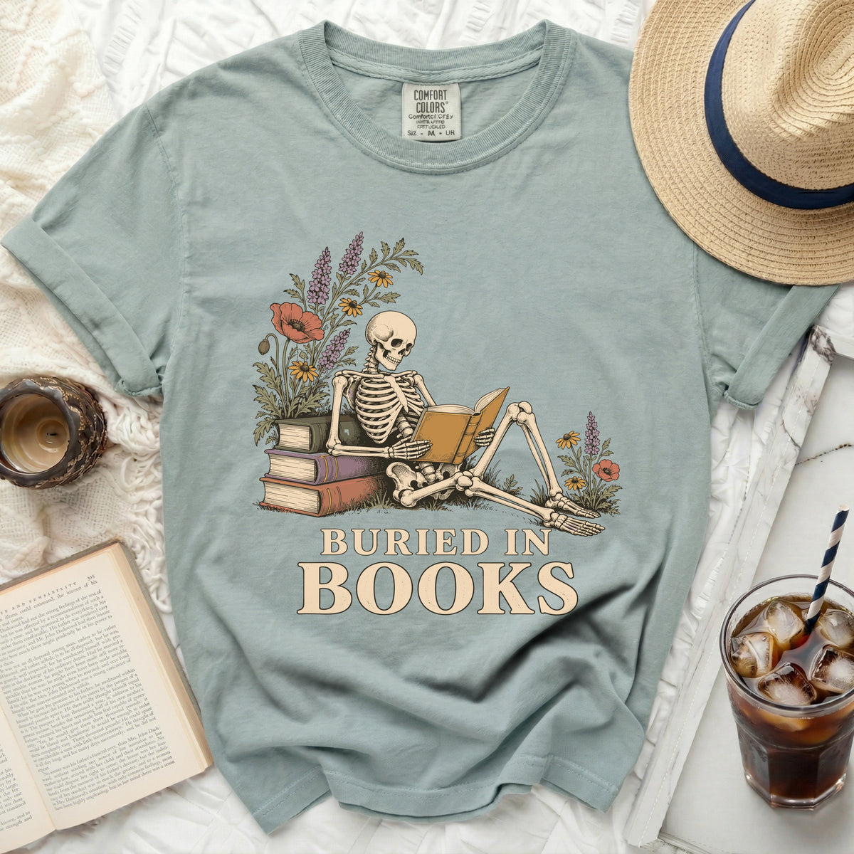Skeleton reading a book with flowers, text "BURIED IN BOOKS" on green t-shirt.