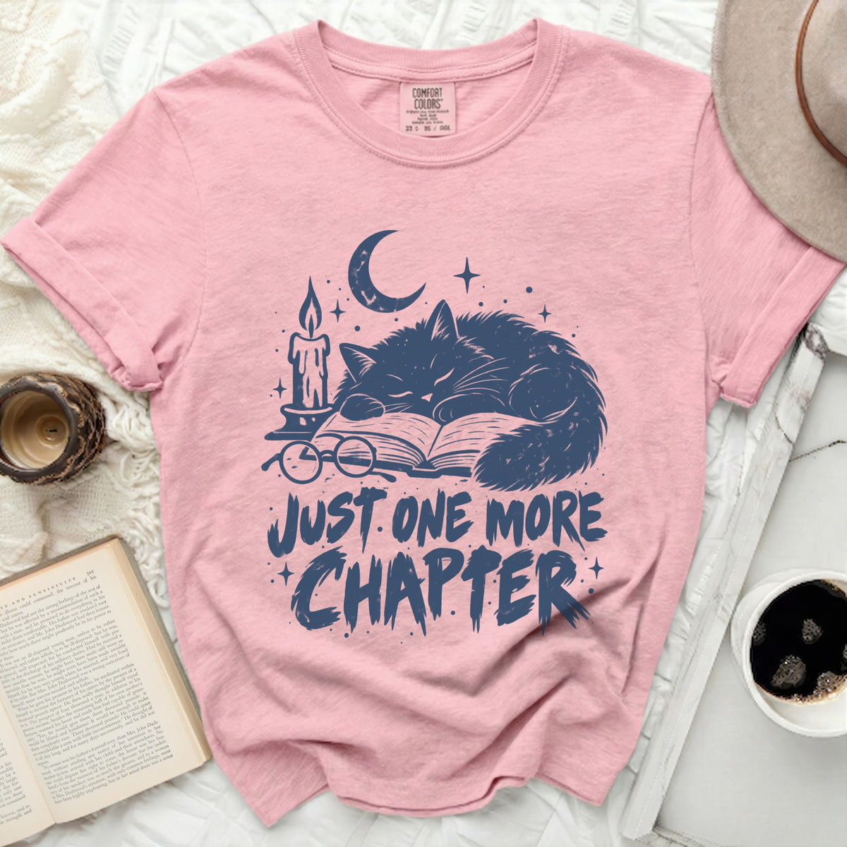 Pink t-shirt with navy graphic of sleeping cat, candle, book, and text "JUST ONE MORE CHAPTER".