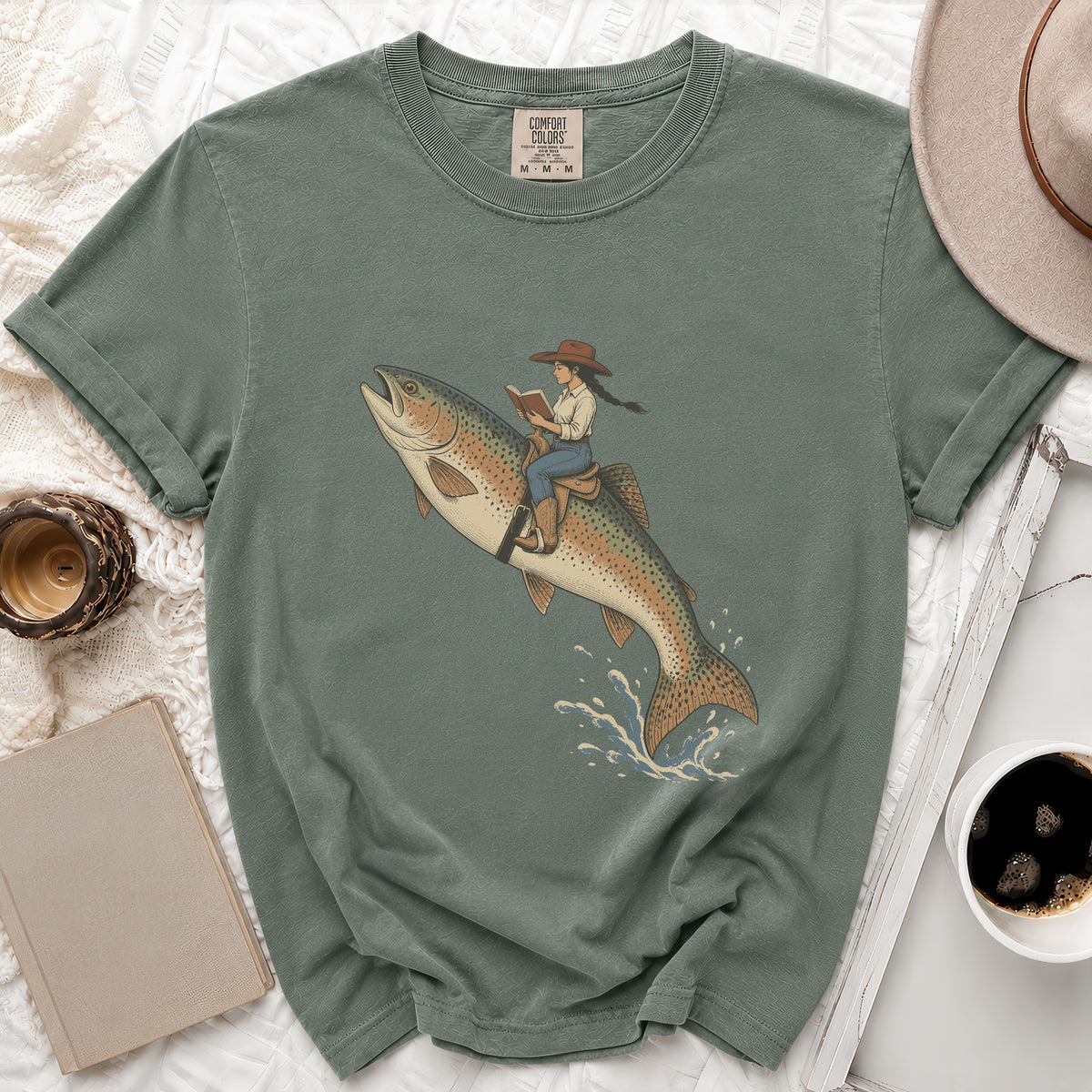 T-shirt with graphic of woman in cowboy hat riding a jumping trout and reading a book.