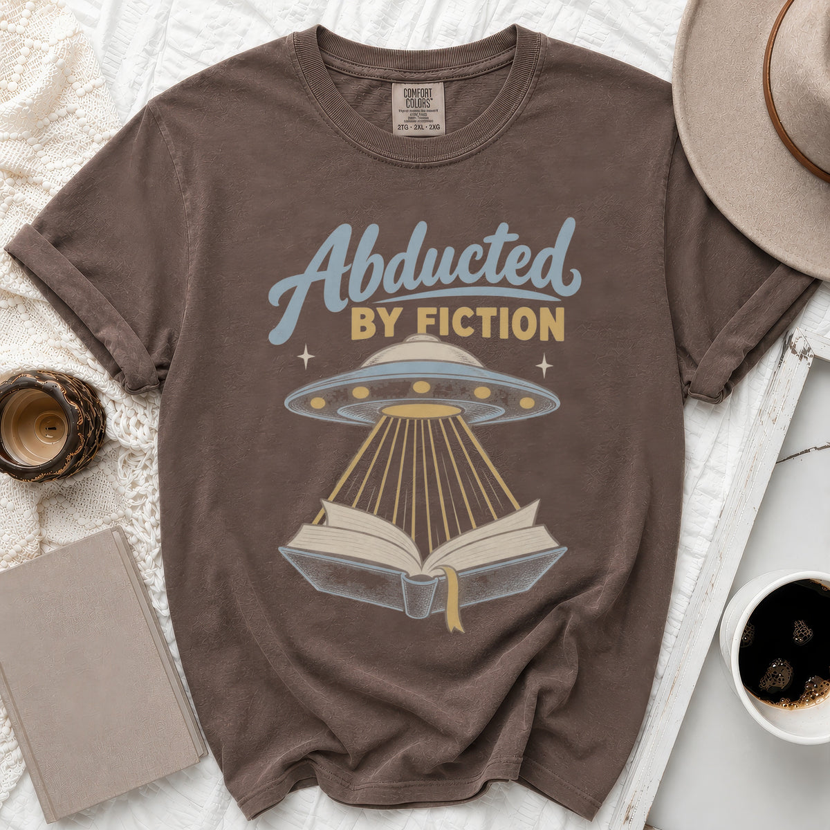 Brown t-shirt with "Abducted by Fiction" text and a UFO beaming light onto an open book.