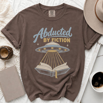 Brown t-shirt with "Abducted by Fiction" text and a UFO beaming light onto an open book.