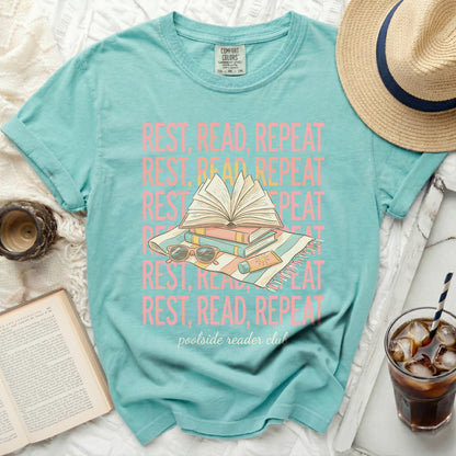 Aqua t-shirt with "REST, READ, REPEAT" text and books on beach towel.