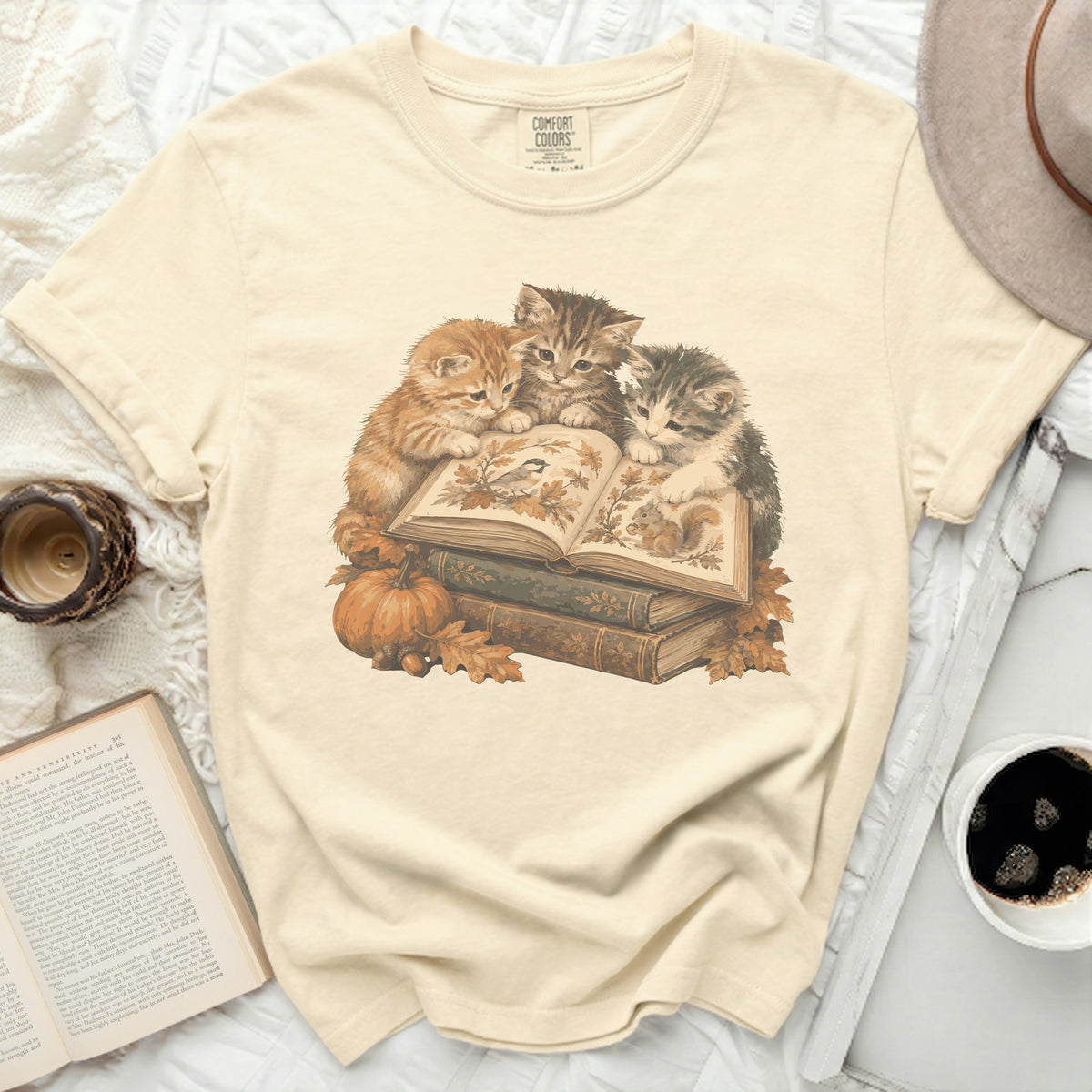 Three kittens look at an open book surrounded by autumn leaves and pumpkins.