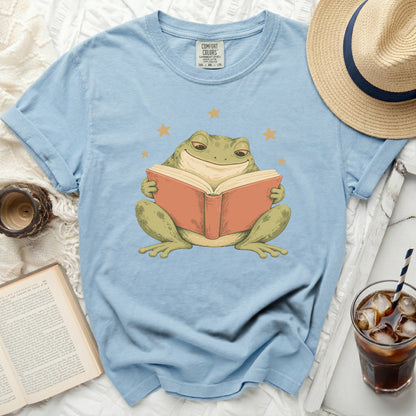 Light blue t-shirt with a graphic of a smiling frog reading a book and stars.