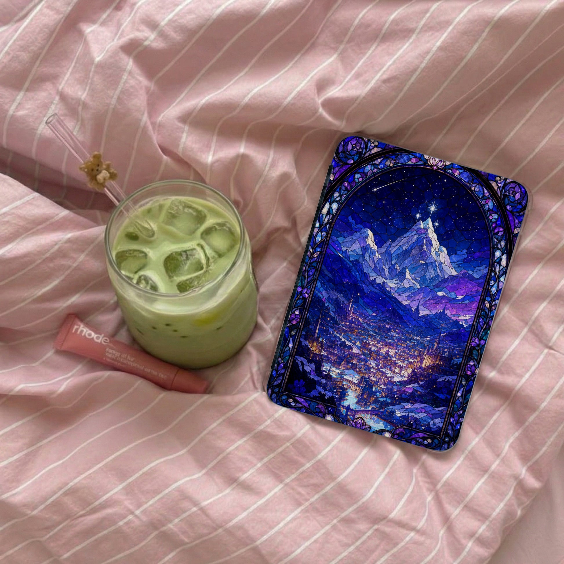 Glass of iced matcha latte next to a cosmetic tube and a stained-glass-style phone case.