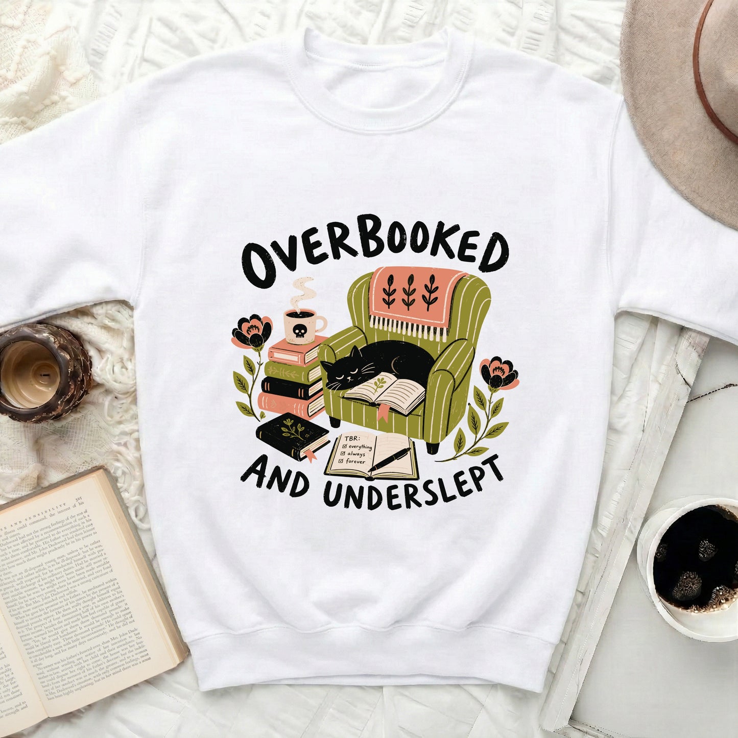 White sweatshirt with black text reading "OVERBOOKED AND UNDERSLEPT" above an illustration.