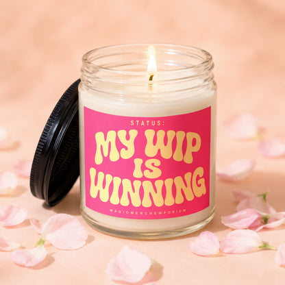 Glass candle with lit wick and pink label that reads "STATUS: MY HIP IS WINNING".