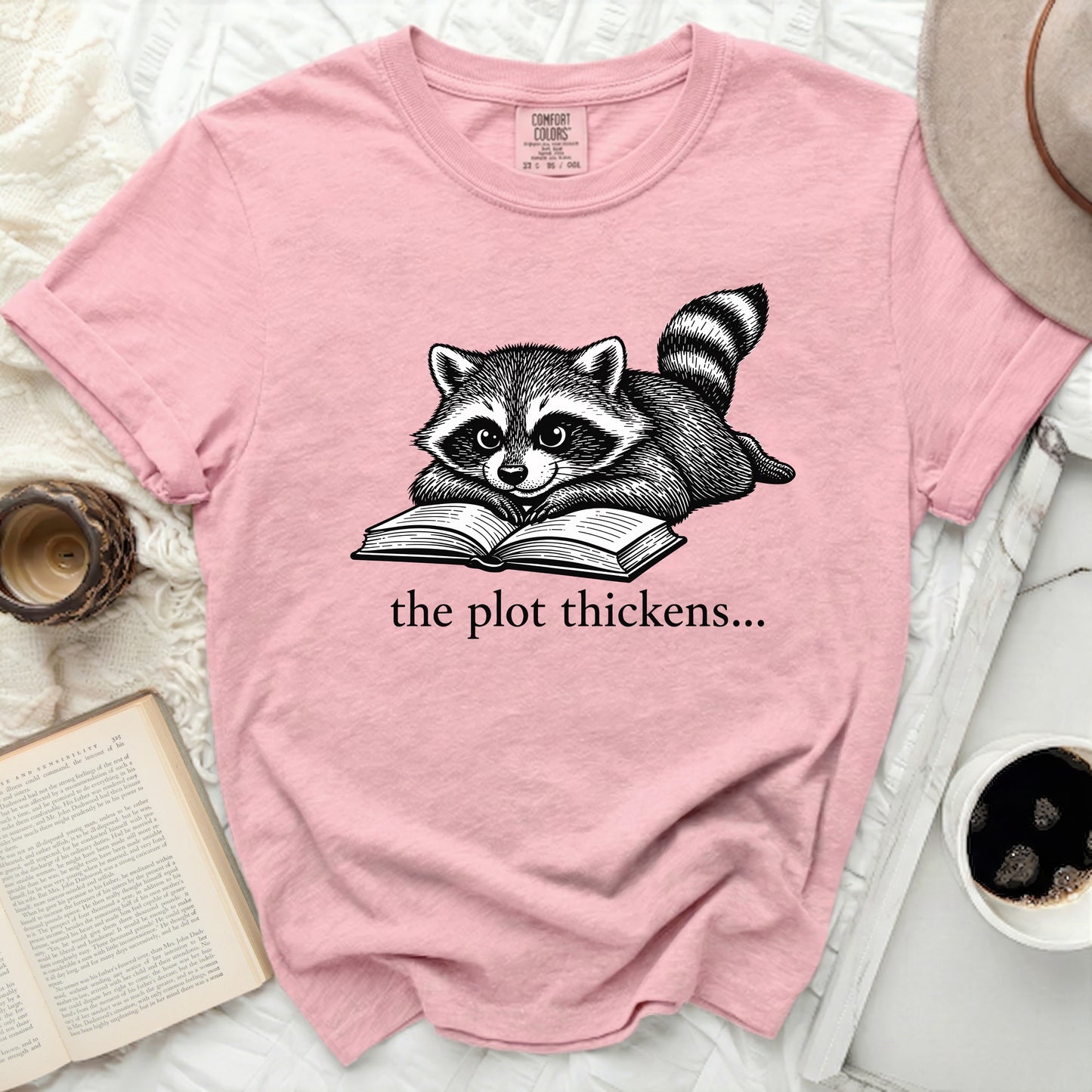 Pink t-shirt with a raccoon reading a book and text "the plot thickens...".